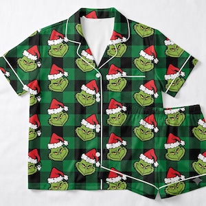 May include: A short-sleeve button-down pajama shirt and matching shorts set featuring a green and black buffalo plaid pattern. The fabric is covered in a repeating print of the Grinch wearing a red and white Santa hat.