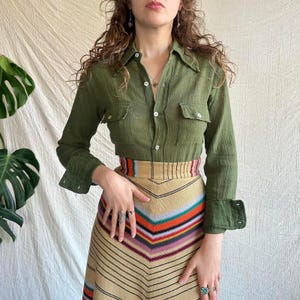 May include: An olive green long-sleeved button-down shirt with two chest pockets and white buttons, paired with a high-waisted skirt featuring a chevron pattern in horizontal stripes of orange, red, blue, purple, and black on a tan background.