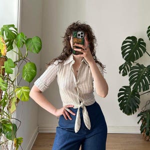 May include: A short-sleeved button-down shirt featuring a cream base with thin vertical stripes in brown and blue. The shirt has a pointed collar and a tie-front hem detail. It is paired with dark blue trousers that have a visible button and zipper.