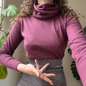 May include: A long-sleeved, plum-coloured turtleneck top made of a soft, knit fabric. The garment features a high, folded collar and a fitted silhouette that extends to the waist, where it is tucked into a metallic, horizontally striped skirt.