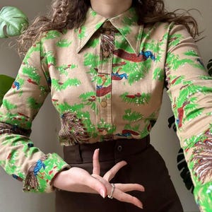 May include: A long-sleeved button-down shirt in a tan hue featuring a repeating pattern of green grass and blue and red peacocks. The shirt has a pointed collar and a central placket with light brown buttons. The fabric displays a detailed print.