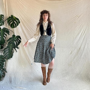 May include: A cream-colored long-sleeve button-down shirt, a dark navy blue velvet vest with a front tie closure, and a dark green midi skirt featuring a small white floral pattern, paired with brown leather lace-up boots and round glasses.
