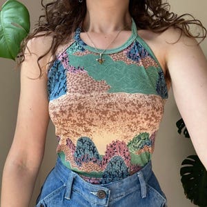 May include: A halter top featuring a landscape print with rolling hills, trees, and a dotted texture in shades of green, blue, pink, and tan. The garment has a high neckline and is paired with blue denim jeans featuring a visible metal button.
