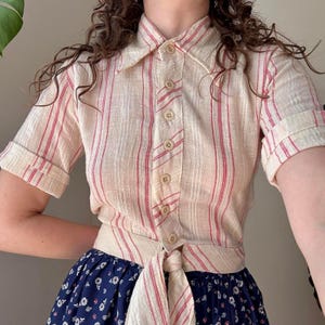 May include: A short-sleeved button-down shirt in a cream-colored fabric with vertical red and white stripes. The shirt features a pointed collar, cuffed sleeves, and a matching fabric tie at the waist, paired with a dark blue skirt with a white floral print.