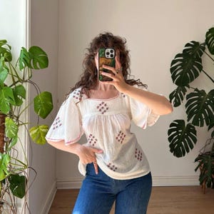 May include: A white blouse with short flutter sleeves and a square neckline. The garment features small, embroidered red diamond-shaped patterns across the front and sleeves, with a delicate red stitched trim along the neckline and sleeve edges.