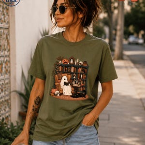 Könnte beinhalten: Ein olivgrünes Kurzarm-T-Shirt mit einem Grafikdruck, der einen Geist zeigt, der in einem Sessel sitzt und ein Buch vor einem Bücherregal liest, zusammen mit einem Kürbis, einem Totenkopf und einer schwarzen Katze auf einem Teppich.