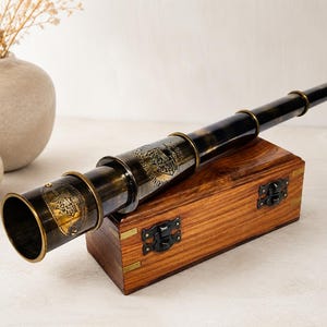 Peut inclure: Une longue-vue nautique en laiton et métal foncé avec un emblème gravé et le texte Marine Telescope 1885. La longue-vue est posée sur une boîte de rangement rectangulaire en bois dotée de loquets en métal noir et de coins en laiton.