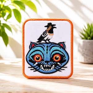 Puede incluir: Un parche bordado cuadrado con borde naranja sobre fondo blanco. El diseño presenta una cabeza de gato azul con grandes ojos naranjas y dientes blancos afilados, coronada por un pájaro negro con sombrero y collar de cadena dorada.