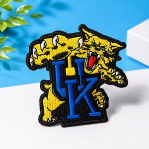 Puede incluir: Un parche bordado que presenta una mascota de gato montés amarilla con rayas negras, dientes blancos y una lengua roja, situada sobre un gran logotipo azul de letras mayúsculas UK. Todo el diseño está delineado con hilo negro y acabado texturizado.