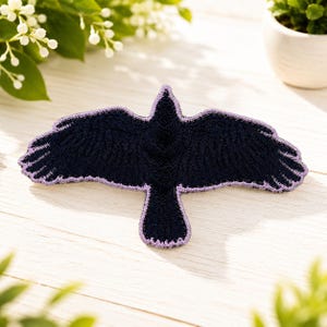 May include: A dark navy blue embroidered patch in the shape of a bird with its wings spread wide. The bird features detailed stitching to represent feathers and is outlined with a thin, light purple border along the entire edge of the shape.