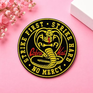May include: A circular embroidered patch featuring a black background with a yellow cobra snake graphic in the center. The text "STRIKE FIRST STRIKE HARD NO MERCY" is embroidered in yellow around the border, with "Cobra Kai" in red script.