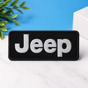 Pode incluir: Um emblema retangular bordado com fundo preto e a palavra Jeep costurada em fio branco. O emblema possui uma borda com acabamento limpo e uma superfície texturizada, exibindo o nome da marca em uma fonte em negrito e bem legível.