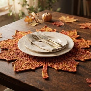 May include: A crochet placemat shaped like a large maple leaf in variegated shades of orange, red, and yellow. Centered on the placemat is a white ceramic plate with a textured rim, a folded beige cloth napkin, a silver fork, and a silver knife.