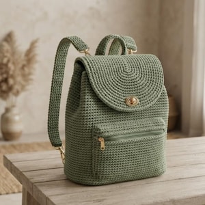 Puede incluir: Una mochila de ganchillo color verde salvia que presenta una solapa superior redondeada con un cierre de giro de metal dorado, un asa superior, correas ajustables con herrajes dorados y un bolsillo frontal con cremallera del mismo tono.