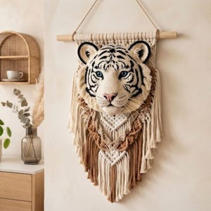 May include: A wall hanging featuring a crocheted white tiger head with black stripes and blue eyes, mounted on a macrame base made of cream and tan cotton cord. The piece is suspended from a horizontal wooden dowel with a thin rope hanger.