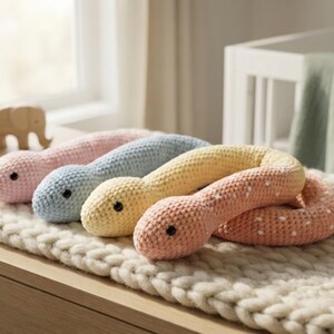 May include: Four long, soft, crocheted snake-shaped toys in pastel pink, light blue, pale yellow, and a coral-orange shade with small white spots. Each toy features a rounded head with two small, dark, circular eyes and a long, flexible body.