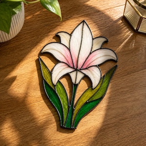 May include: A stained glass suncatcher shaped like a lily flower with white and soft pink petals, accented by vibrant green and yellow leaves. The piece is constructed with dark metal solder lines outlining each individual glass segment.