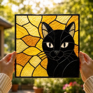 May include: A square stained glass panel featuring a black cat silhouette centered against a vibrant yellow and gold sunburst background. The cat has white glass eyes and is outlined in black metal cames, creating a geometric mosaic effect.