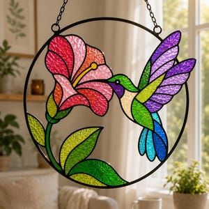 Può includere: Un acchiappasole circolare in vetro colorato raffigurante un colibrì con ali e corpo in vetro testurizzato verde, viola e blu, vicino a un grande fiore di ibisco rosa e rosso con foglie verdi, il tutto racchiuso in una cornice di metallo nero.