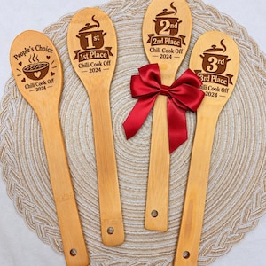 May include: Four light-colored wooden spoons engraved with Chili Cook Off 2024 designs. The spoons feature text for People's Choice, 1st Place, 2nd Place, and 3rd Place. A dark red satin ribbon is tied around the 2nd place spoon handle.