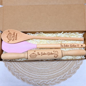 May include: A set of three kitchen utensils in a cardboard box, including a wooden spoon, a light pink silicone spatula with a wooden handle, and a wooden rolling pin. Each item is engraved with the text The Baker Kitchen and pie icons.
