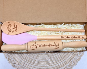 May include: A set of three kitchen utensils in a cardboard box, including a wooden spoon, a light pink silicone spatula with a wooden handle, and a wooden rolling pin. Each item is engraved with the text The Baker Kitchen and pie icons.