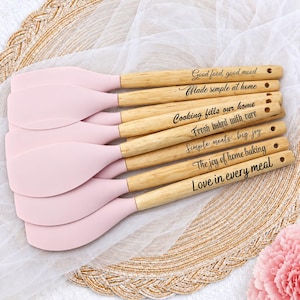 May include: A set of six kitchen spatulas with light pink silicone heads and light-colored wooden handles. Each handle features black engraved text: Good food, good mood, Made simple at home, Cooking fills our home, Fresh baked with care, Simple meals, big joy, The joy of home baking, and Love in every meal.