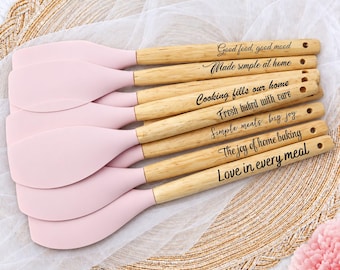 May include: A set of six kitchen spatulas with light pink silicone heads and light-coloured wooden handles. Each handle features black engraved text: Good food, good mood, Made simple at home, Cooking fills our home, Fresh baked with care, Simple meals, big joy, The joy of home baking, and Love in every meal.