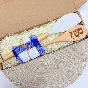 May include: A kitchen set featuring a white silicone spatula and a wooden spoon with the engraved name Blake and a floral letter B. The utensils are wrapped in a blue and white checkered cloth with a thin brown string, nestled in a cardboard box.