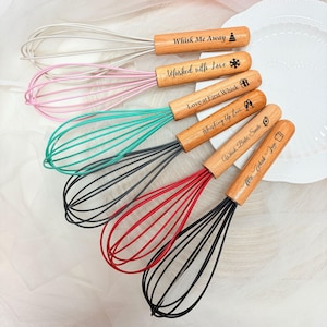 May include: A set of six kitchen whisks with light wood handles and colourful wire loops in white, pink, teal, dark grey, red, and black. Each handle features engraved text and icons, including Whisk Me Away, Whisked with Love, Love at First Whisk, Whisking Up Love, Whisk, Bake, Smile, and Mix, Whisk, Love.