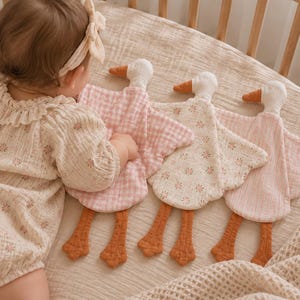 May include: Three soft plush goose security blankets with long orange legs and feet. One features a pink and white gingham pattern, one has a cream fabric with a small floral print, and the third is a solid light pink, all with white heads.