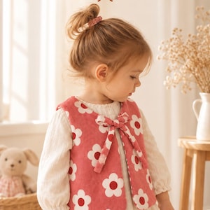 Könnte beinhalten: Eine rosafarbene Cordweste mit einem weißen und roten Blumenmuster und einer Bindeschleife vorne. Der Text "Vest Pattern" steht oben im Bild. Die Weste wird über einem cremefarbenen Kleid mit langen Ärmeln und gerafften Bündchen getragen.
