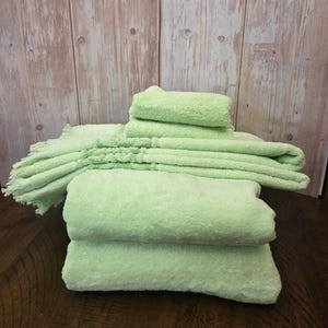 May include: A stack of light green towels featuring a soft, textured terry cloth surface. The set includes multiple folded towels of varying sizes, with one towel displaying a decorative fringe detail along its edge, creating a cohesive collection.