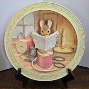 May include: A round decorative plate featuring a three-dimensional relief of a mouse wearing glasses and reading a newspaper titled The Tailor. The mouse sits on a pink spool of thread surrounded by sewing tools, a candle, and a floral border.