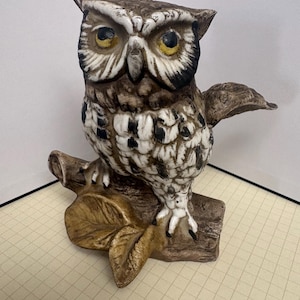 May include: A ceramic owl figurine perched on a textured branch. The owl features brown, white, and black plumage with prominent yellow eyes and dark pupils. The branch is brown with a leaf detail at the base, crafted with a glossy finish.