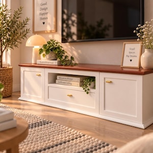 Può includere: Un mobile porta TV bianco con un piano in legno marrone, dotato di due armadietti laterali e un ripiano centrale aperto con un cassetto. Ogni sezione ha una piccola maniglia dorata. Il mobile contiene libri intitolati Miniature Homes e Dollhouse Living.