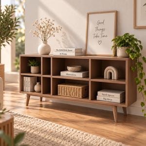 Può includere: Un mobile porta TV in legno marrone chiaro con diversi scomparti rettangolari, sostenuto da quattro gambe affusolate. L'unità contiene vari oggetti decorativi tra cui libri, un cesto intrecciato, una ciotola e una piccola scultura ad arco.