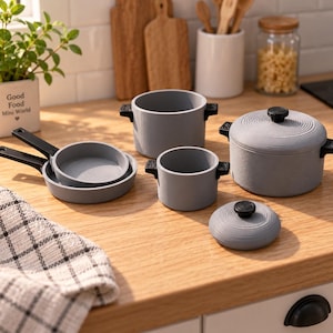 Puede incluir: Un juego de utensilios de cocina en miniatura de color gris que incluye dos sartenes con mangos negros, dos ollas con asas laterales negras y una tapa a juego con un pomo negro. Los artículos tienen un acabado texturizado sobre madera.