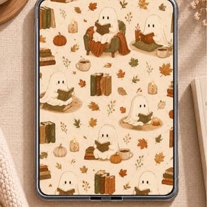 May include: A digital tablet displaying a repeating pattern of white ghosts reading books, surrounded by autumn elements including orange pumpkins, brown fallen leaves, stacks of books, lit candles, and mugs of coffee on a light beige background.