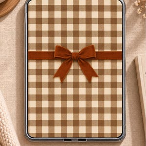 May include: A rectangular tablet device featuring a clear protective case and a screen display with a brown and cream-colored gingham check pattern. A horizontal brown velvet ribbon with a centered bow is placed across the middle of the screen.