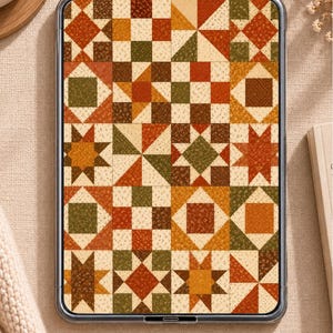 May include: A digital tablet displaying a quilt pattern featuring a grid of squares with geometric designs in shades of orange, brown, cream, and olive green. The patterns include star shapes, pinwheels, and checkerboard motifs with textured fabric prints.