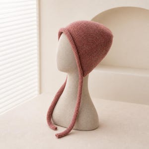 May include: A dusty rose knitted bonnet featuring a smooth, rounded crown and a rolled edge along the front opening. Two long, matching knitted ties extend from the sides of the bonnet, hanging down to secure the headpiece under the chin.