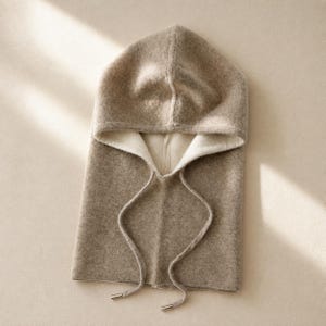 May include: A beige hooded balaclava featuring a soft, textured knit construction. The hood includes a wide opening with a cream-colored interior lining and two long, matching beige drawstrings that hang down, finished with small metallic silver end caps.