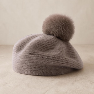 May include: A light taupe knit beret featuring a ribbed texture throughout the crown and a large, fluffy, matching faux fur pom-pom attached to the top center. The hat is constructed from a soft, fuzzy yarn material with a rounded, classic shape.
