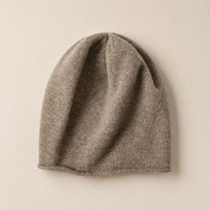 May include: A solid light brown beanie hat with a soft, fine-knit texture. The headwear features a rounded crown and a simple, unadorned rolled edge at the base, showcasing a consistent, neutral earth-toned color throughout the entire garment.