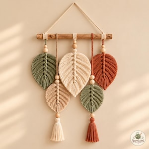 May include: A macrame wall hanging featuring five leaf-shaped fibre art pieces in olive green, cream, and terracotta colours. The leaves are suspended from a horizontal wooden branch with a rope hanger, accented by natural wooden beads and tassels.