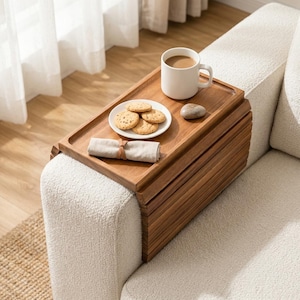 May include: A rectangular wooden armrest tray with a flexible slatted design draped over a cream-colored sofa arm. The tray holds a white ceramic mug filled with coffee, a small white plate with four round cookies, a grey stone, and a folded beige napkin.