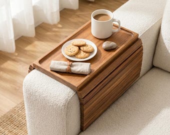 May include: A rectangular wooden armrest tray with a flexible slatted design draped over a cream-colored sofa arm. The tray holds a white ceramic mug filled with coffee, a small white plate with four round cookies, a grey stone, and a folded beige napkin.