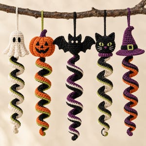 Pode incluir: Cinco enfeites de Halloween em crochê, incluindo um fantasma branco, uma abóbora laranja, um morcego preto, um gato preto e um chapéu de bruxa roxo. Cada figura está presa a uma longa cauda de fio em espiral multicolorida pendurada num ramo.