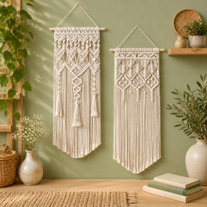 May include: Two off-white macrame wall hangings suspended from wooden dowels with white string. Each piece features intricate knotted patterns, including diamond shapes and vertical fringe, creating a textured, layered appearance on the wall.